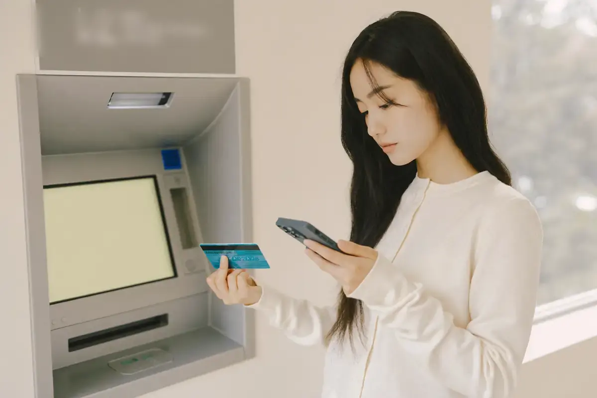 은행 ATM 앞에 선 여성이 한 손에는 카드를, 다른 손에는 스마트폰을 들고 화면을 보고 있다. ATM 화면은 비어 있다.