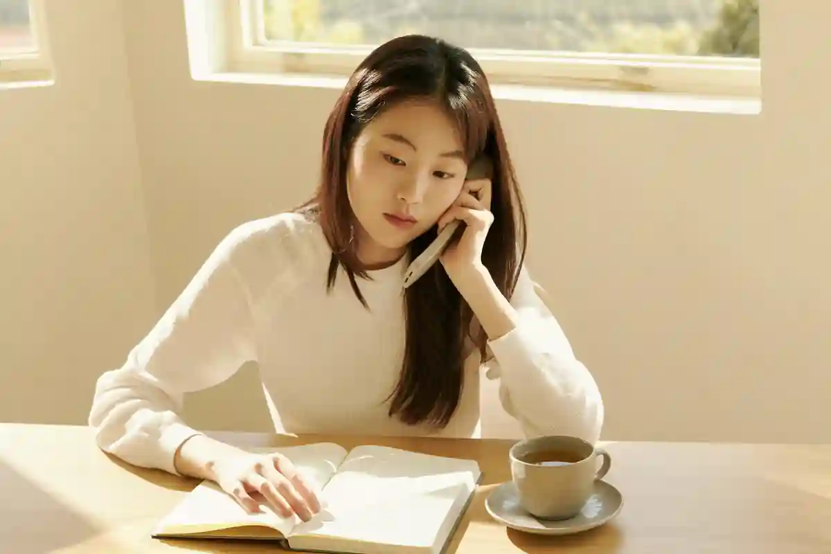 창가 책상에 앉아 한 손에 전화기를 들고 통화하는 여성. 다른 손은 덮인 빈 노트 위에 놓여 있고 옆에 찻잔이 있다.