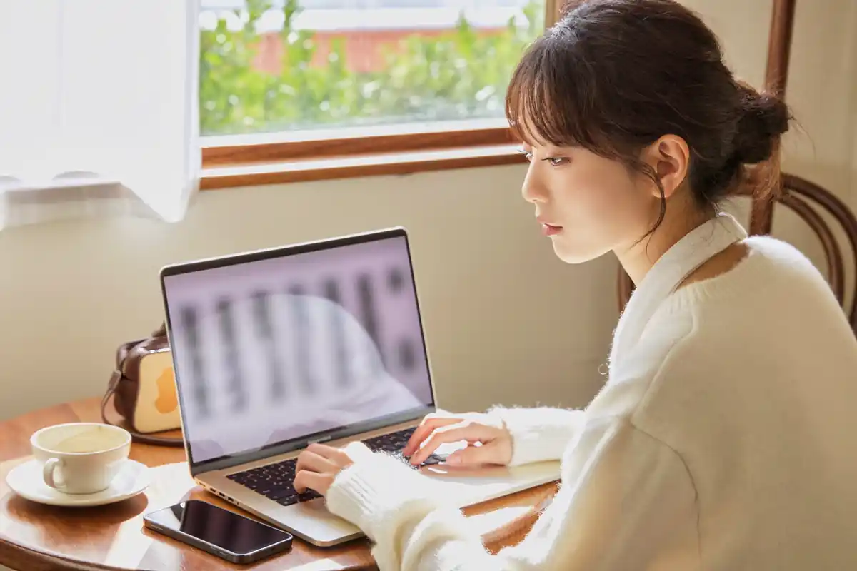 창가 자리에 앉아 노트북으로 무언가를 입력하는 여성, 옆에 커피잔과 엎어 둔 휴대전화가 있다.