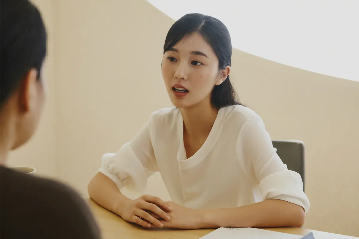 탁자를 사이에 두고 마주 앉아 말하고 있는 여성, 맞은편 사람은 뒷모습만 보인다.