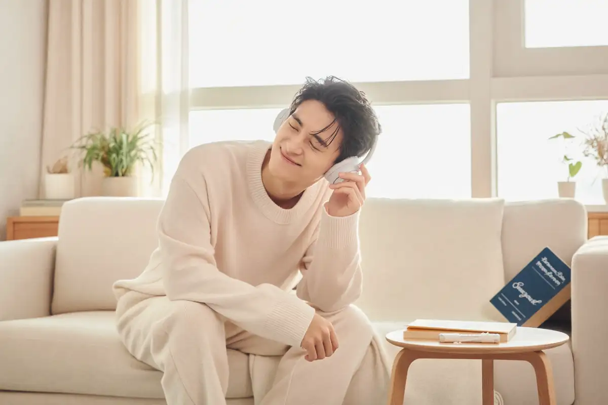 볕이 드는 거실 소파에서 흰 헤드폰을 쓰고 눈을 감은 채 듣고 있는 남성.