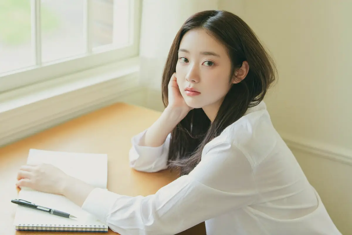 창가 나무 책상에 앉아 턱을 괴고 생각에 잠긴 여성, 앞에는 펼쳐 둔 공책과 볼펜.