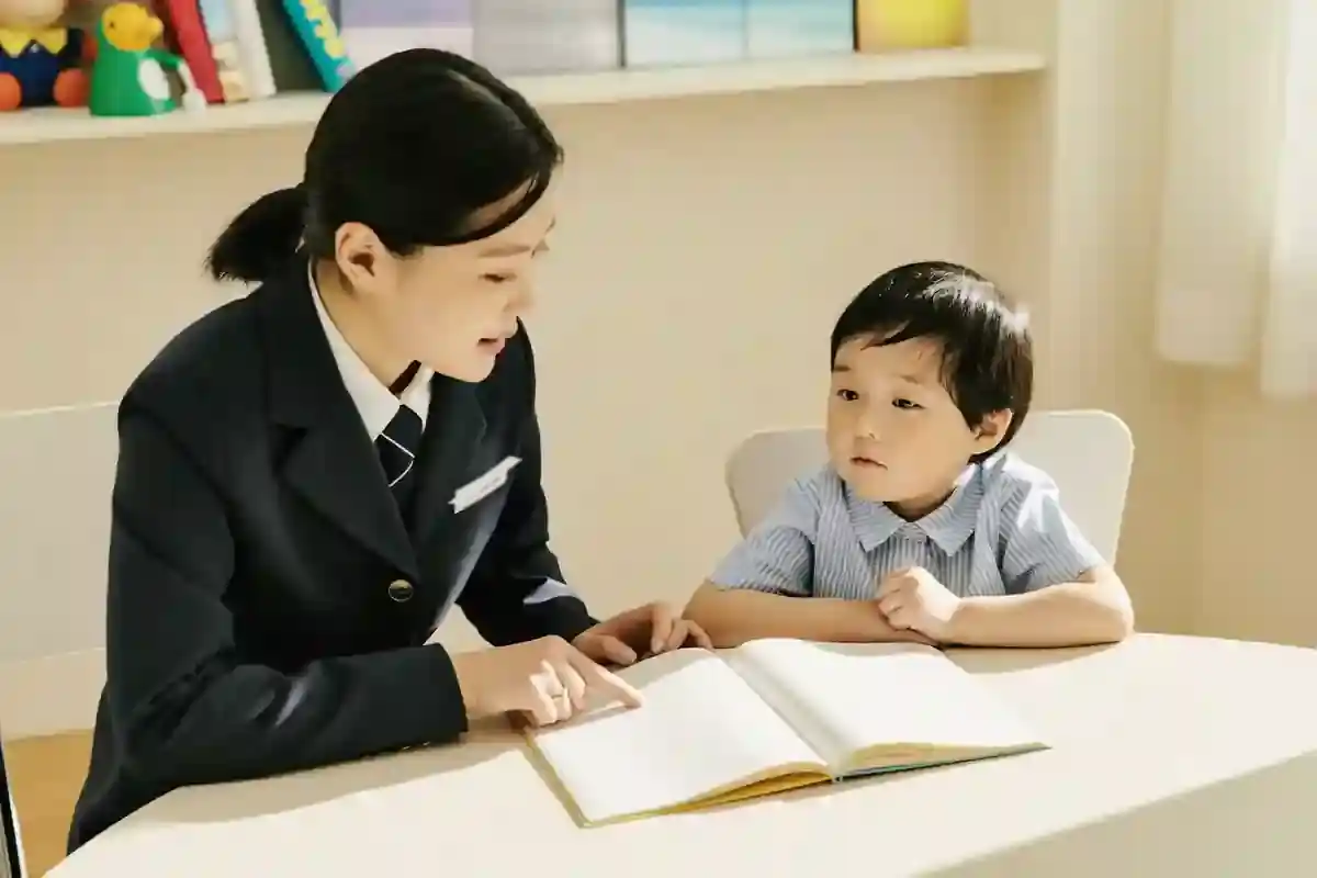 작은 탁자에 마주 앉은 교사와 아이가 펼쳐진 빈 공책을 보며 이야기하고 있다.