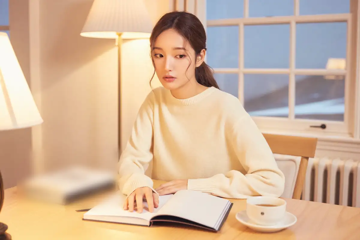 저녁 식탁에서 스탠드 불빛 아래 펼친 공책에 손을 얹고 혼자 되뇌는 여성. 옆에 찻잔이 놓여 있다.