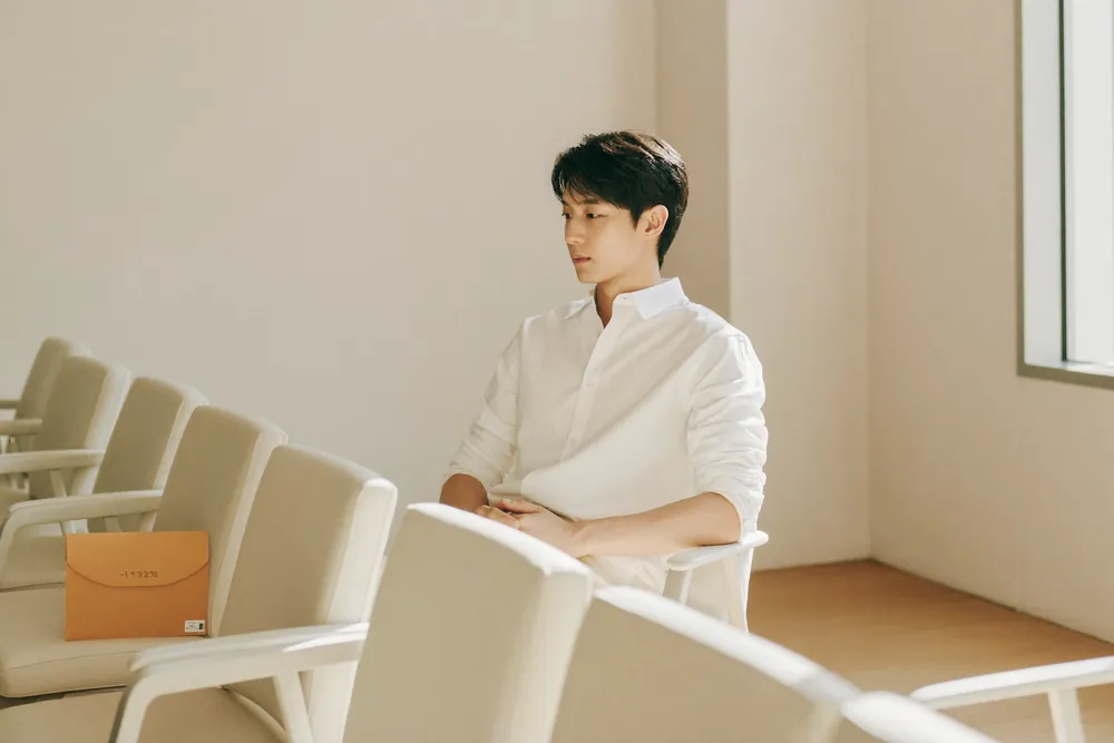 볕이 드는 대기실에서 밝은 의자에 앉아 차분히 기다리는 남성. 옆자리에 서류 봉투가 놓여 있다.