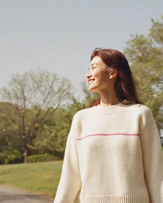 아침 볕이 드는 강변 공원 산책로를 걷는 여성. 뒤로 풀밭과 나무, 맑은 하늘이 펼쳐진다.